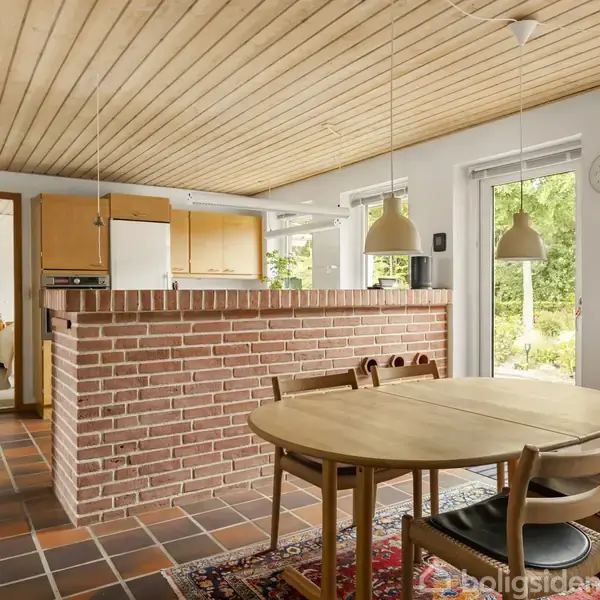 Et rundt træbord med fire stole står på et tæppe. En murstensbar deler køkkenet med lyse trælofter, mens store vinduer lukker udsyn til en frodig have ind.