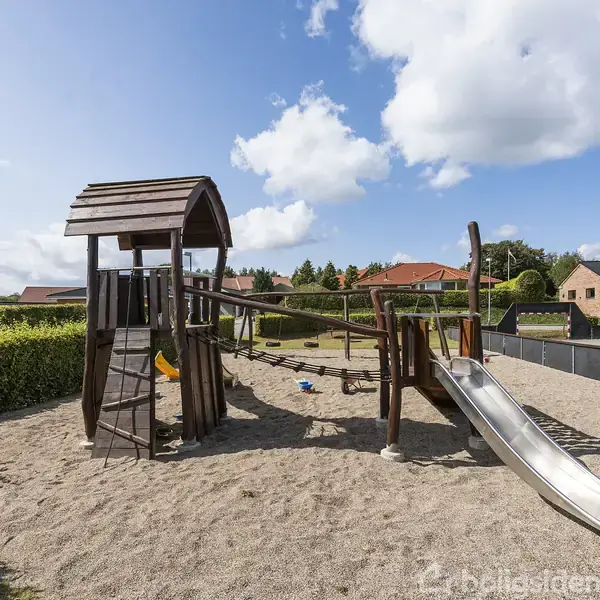 Legetårn af træ med rutsjebane står i en sandkasse på en legeplads. Det er omgivet af grønne hække og boliger i baggrunden under en blå himmel med skyer.