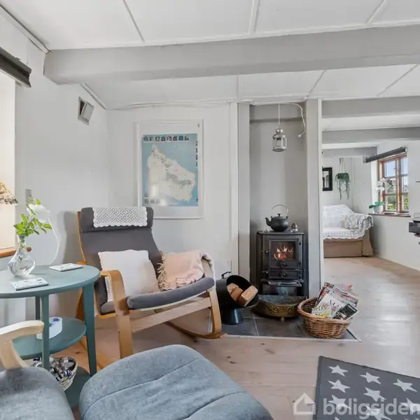 En hyggelig stue har en brændeovn under et vindue med gardiner. Ved siden af står en grå lænestol med puder og et lille rundt bord. En kurv med aviser er ved siden af ovnen.