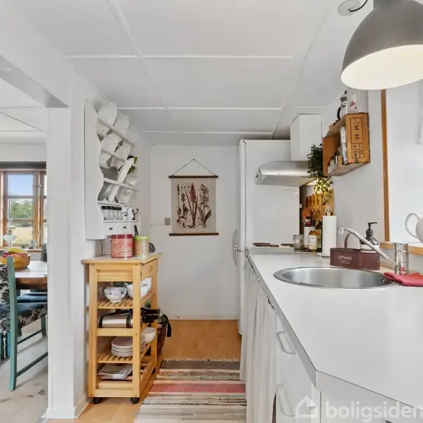 Et køkken med hvide skabe og bordplade, vaskeplads og diverse køkkenredskaber. Ved siden af er en spiseplads med blå mønstrede stole, og store vinduer slipper naturligt lys ind.