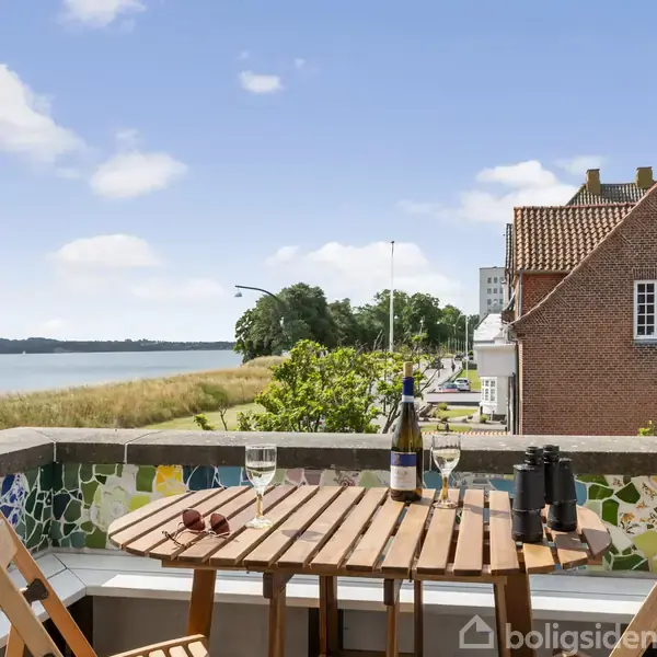 Et træbord står på en balkon med vinflaske, glas, og kikkert. Udsigt mod havnen med huse og grøn natur i baggrunden. Vejret er klart med blå himmel.