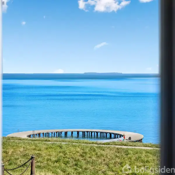 Rund badebro står stille i det klare, blå hav, set gennem et vindue. Grøn græsplæne omgiver broen, med en vid horisont og blå himmel i baggrunden.