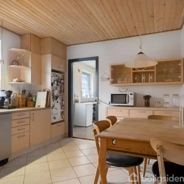 Køkkenbord med stole står i et lyst køkken. Køleskab og opvaskemaskine til venstre, skabe med glas og mikrobølgeovn hænger på væggen. Træloft og flisegulv skaber en hyggelig atmosfære.