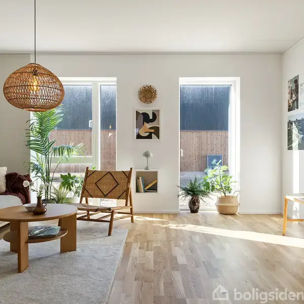 En lys stue med en grå sofa til venstre, hvor der ligger puder. En rund træbord står foran sofaen. En pendellampe hænger over bordet. To store vinduer lukker lys ind, omgivet af grønne planter. En bænk og vægkunst findes til højre.