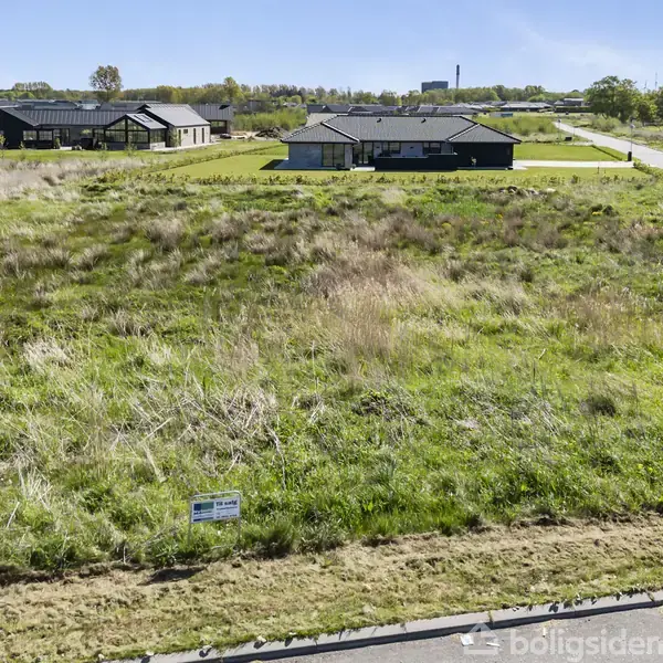 Græsmark med et skilt i forgrunden, omgivet af nybyggede huse. Skiltet lyder: "GRUND TIL SALG EDC Mæglerne." I baggrunden ses træer og en klar blå himmel.