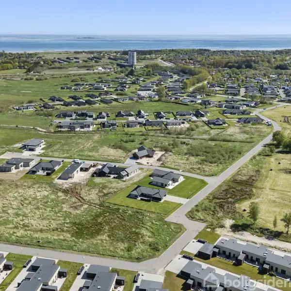 Huse, placeret strategisk i et grønt boligområde, ligger spredt i en forstadslignende scene. I baggrunden ses åbne marker og en kystlinje ved horisonten, der skaber en rolig atmosfære.