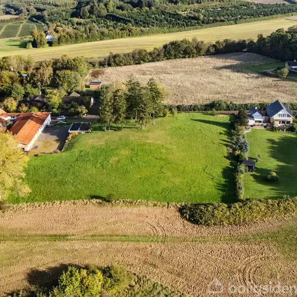 Luftfoto af landdistrikt med flere huse, marker og skove. En stor græsplæne strækker sig midt i billedet omgivet af landbrugsjord og træer.