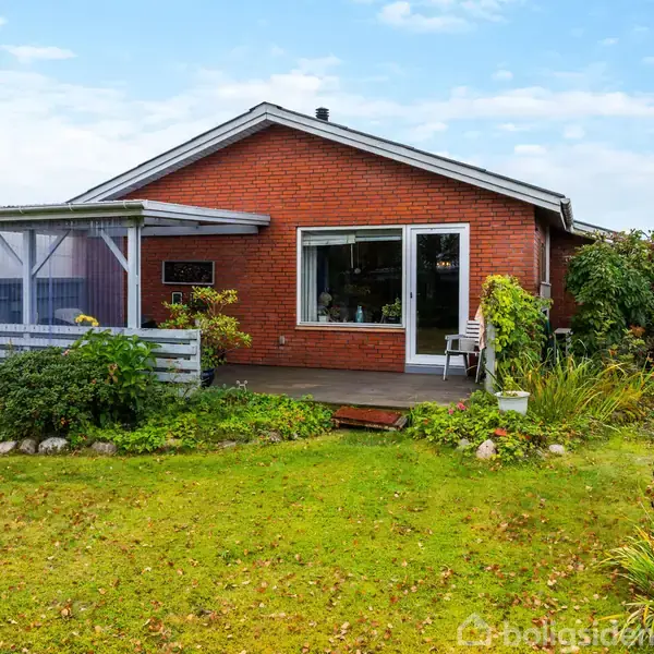 Rødt murstenshus med overdækket terrasse, stående i en velplejet have med græsplæne og buske. En grussti fører til huset, der har store vinduespartier. Skyfri himmel ses i baggrunden.