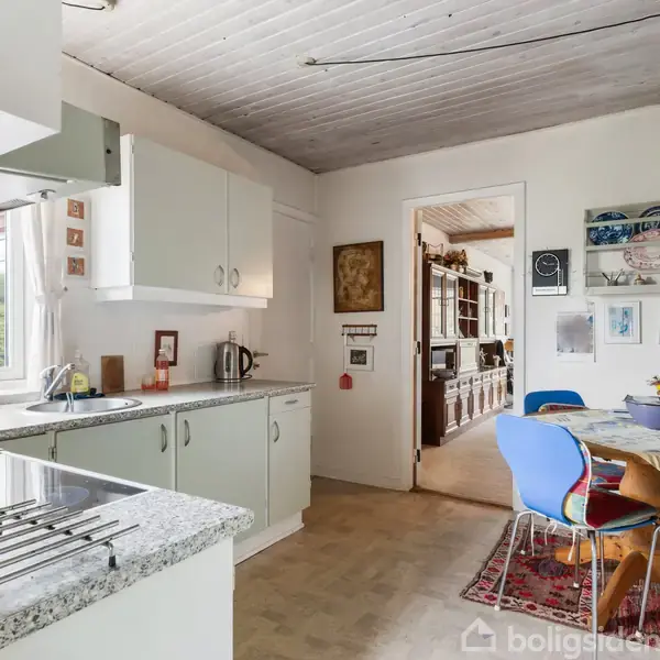 Køkken med lyse skabe (objekt) står stille (handling), mens et bord med farverige stole og dekorerede vægge ses i baggrunden (kontekst).