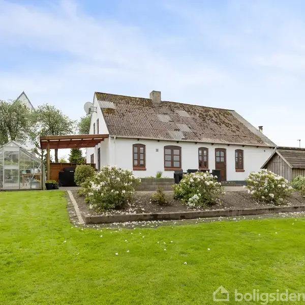 Hvidt hus med mørkt tag står på en velplejet græsplæne, omringet af blomsterbede. Et drivhus ses til venstre, og der er en terrasse med træpergola ved huset.