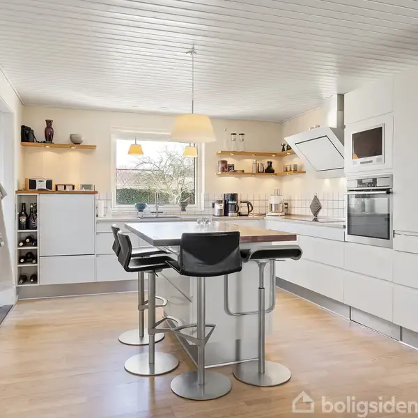 Et moderne køkken med en indbygget ø og barstole, omgivet af hvide skabe og apparater. Vindue med udsigt til træer, overflader med dekorationer, gulv i lyst træ.
