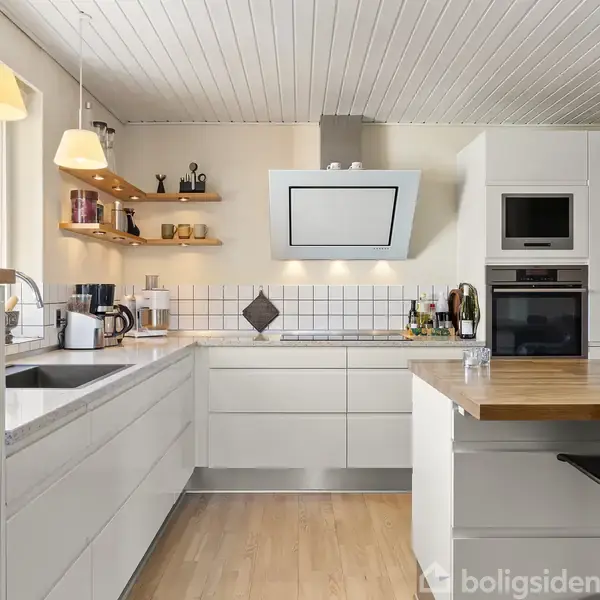 Et moderne køkken med hvide skabe og træbordplade dominerer. Hylder indeholder små dekorative genstande. Ovne er indbygget i en skabsvæg. Hængelamper lyser rummet op.