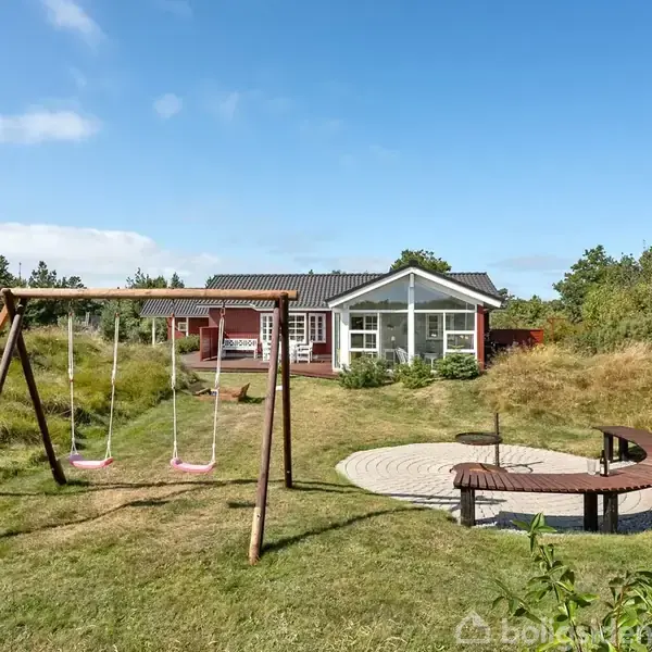 En gyngestativ med to gynger står i en græsplæne. I baggrunden ses et rødt hus med store vinduer omgivet af grønne buske og træer under en klar blå himmel.