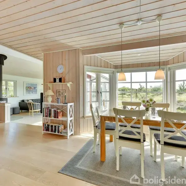 Et spisebord med fire stole står ved et vindue med udsigt til naturen udenfor. Stuen, indrettet med lyse trægulve og vægge, har også en reol og en brændeovn.