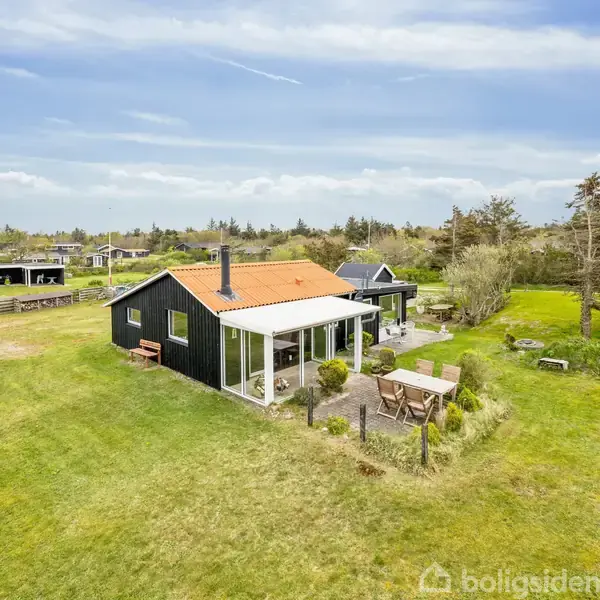 Et sort sommerhus med rødt tag står på en stor græsplæne. Foran huset er der en terrasse med havemøbler. Huset ligger i et naturskønt, åbent område med træer i baggrunden.