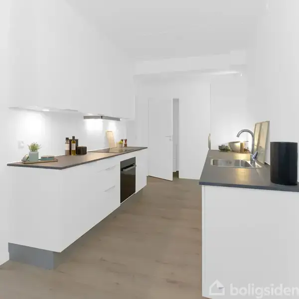 Et moderne køkken; rene, hvide skabe og sorte bordplader; er udstyret med indbygningsovn, kogeplader, og vask; placeret i en lys gang ved siden af en stue med sofa.