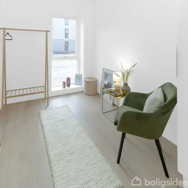 En grøn stol står i et lyst rum ved siden af et lille bord med en lampe og dekorationer. En høj spejl og tøjstativ ses i baggrunden nær et vindue.