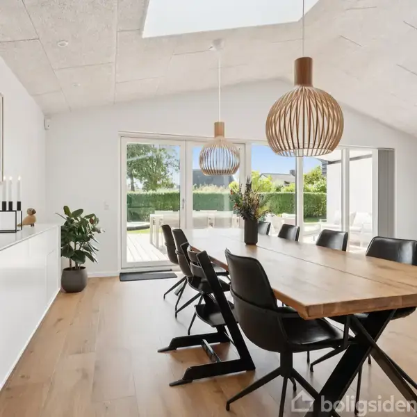 En stor, lys spisestue med et langt træbord omgivet af sorte stole, to moderne lamper hænger over bordet. Store glasdøre fører ud til en terrasse med grøn udsigt.