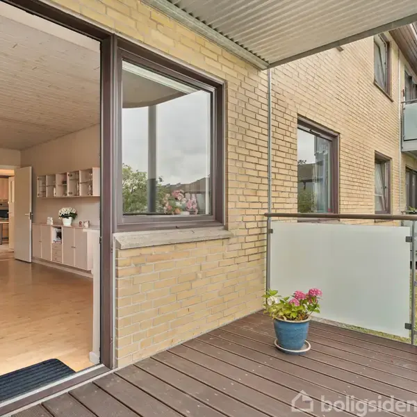 En altan med trægulv har en blå blomsterkrukke. Indgangsdøren står åben til en lys, moderne stue med trægulv. Baggrunden viser et murstensbyggeri med glasbalkon.