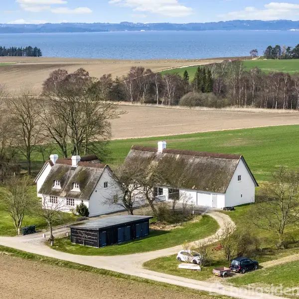 Et stråtækt hus ligger ved en grusvej omgivet af åbne marker og træer, med havet i baggrunden. Der er også køretøjer parkeret i gården.