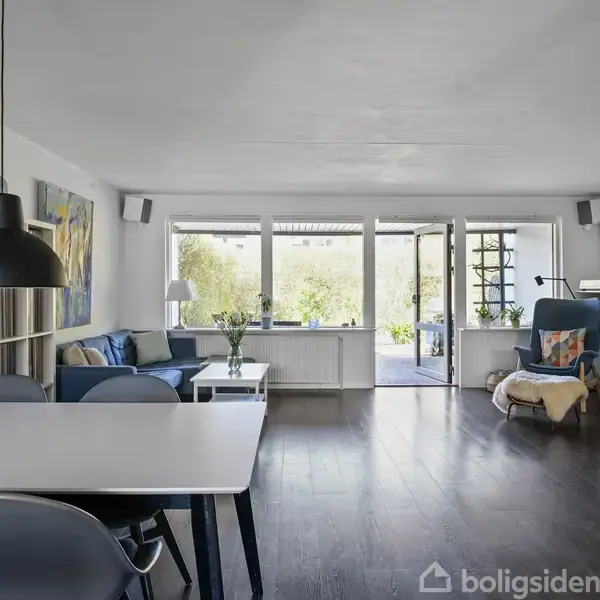 Spisestuemøbler dominerer rummet med et stort bord og stole, mens en sofa og en lænestol er placeret ved vinduerne, der åbner ud til en terrasse med udsigt til grønne omgivelser.