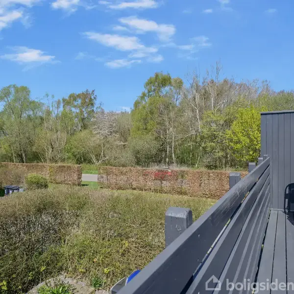 En terrasse med en hvilestol står stille, omgivet af hække og træer i en grøn have. Skuret og blå himmel optræder i baggrunden, og det er en rolig dag.