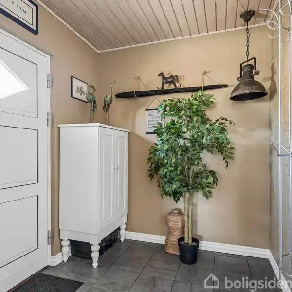 Hvidt skab står i et vindfang med beige vægge. En grøn plante i potte står ved siden af. Hylde på væggen med figurer og lampe hænger fra loftet. Skilt på væggen: "Welcome".