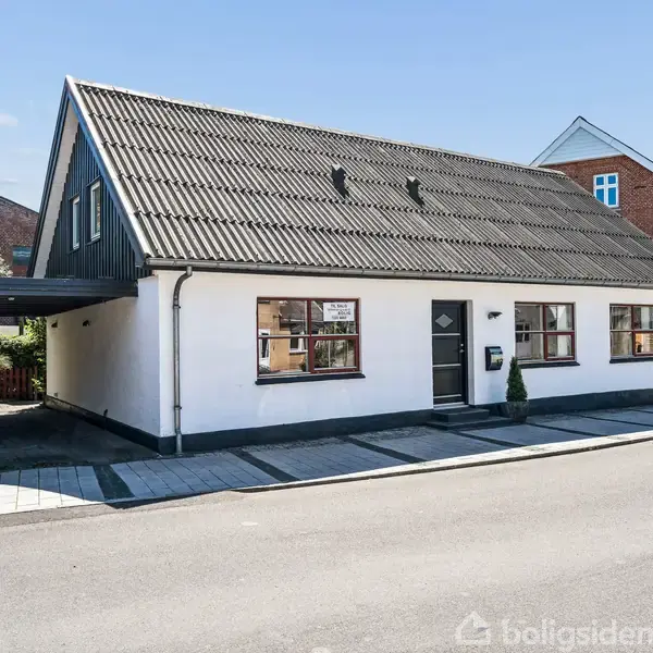 Et hvidt hus med sort tag ligger ved en vej, omkranset af andre bygninger. Huset har vinduer og en dør med et lille træ ved siden af.
