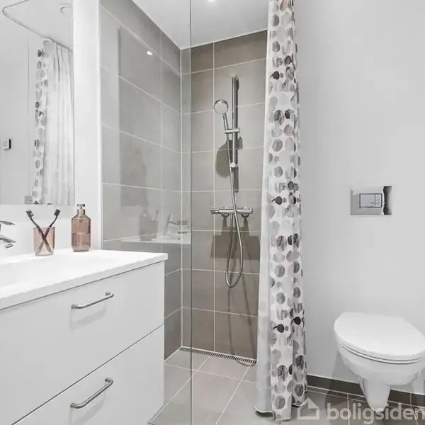 Et moderne badeværelse med en bruser i glasafdækning, en hvid håndvask og skabe. Toilet til højre og en dekorativ bruseforhæng med mønster. Miljøet er lyst og pænt.