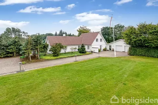 Et hvidt hus med rødt tag står i en velplejet have med grøn græsplæne. En flisebelagt indkørsel fører til en garage. Træer omgiver ejendommen under en blå himmel.
