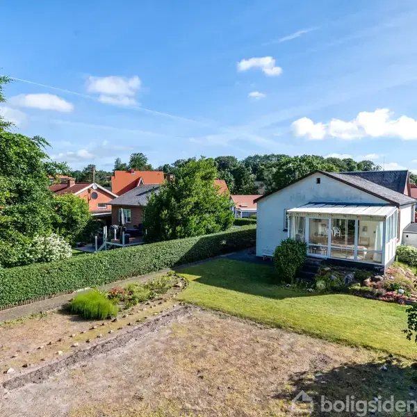 En hvid villa med glasudestue står i en have med græsplæne og buske. Omkring huset ses flere huse og træer under en blå himmel med skyer.