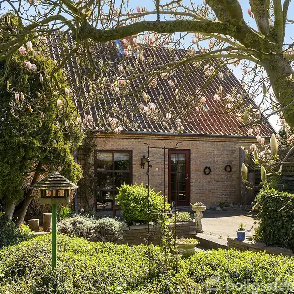 Et murstenshus med skråt tag omgivet af en frodig have. Blomstrende træer og buske skaber en hyggelig atmosfære. En terrasse med krukker og møbler er synlig foran huset.
