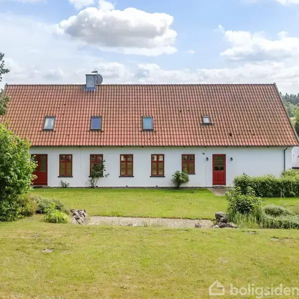 Et hvidt hus med rødt tag og røde døre står på en velplejet græsplæne omgivet af buske og træer. Huset har flere kvistvinduer og ligger i et naturskønt område.