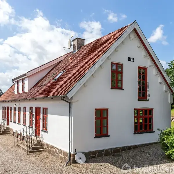 Et hvidt hus med rødt tegltag og røde vinduesrammer står i en grusbelagt have. Datoen "1875" er malet på gavlen. Landskabet er grønt med træer.