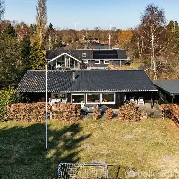 Et sorttagshus står i en have med græsplæne og hæk. Huset har en terrasse med møbler, og en garage er til højre. Omkringliggende træer giver en skovagtig stemning.