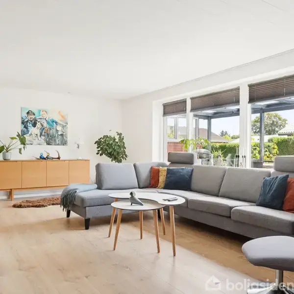 Sofa i grå farve står ved siden af et sofabord; placeret i en lys stue med store vinduer, træmøbler, grønne planter og farverige puder i et moderne hjemmemiljø.