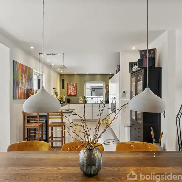 Et moderne køkken-alrum med træbord og stilfulde lamper. Orange stole omgiver bordet, og en mørk skænk står mod bagvæggen. Kunstværker hænger på de hvide vægge.