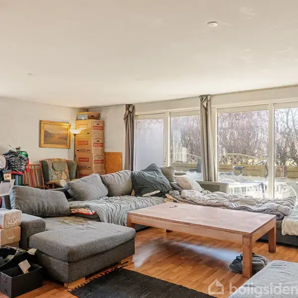 En stor stue med en grå sofa i hjørneformation. Sofabord står foran sofaen. Rummet er fyldt med flyttekasser, småborde og udsigt til en have gennem store vinduer.