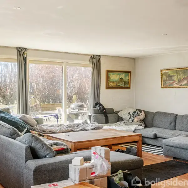 Stor grå hjørnesofa står i en stue med træbord, omgivet af lyse vægge og malerier. Store vinduer giver udsigt til en terrasse og buske udenfor.