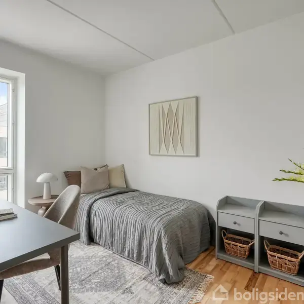 En enkeltseng dækket med gråt sengetæppe står i et lyst værelse. Et skrivebord med stol er ved vinduet, dekoration hænger på væggen, og to grå kommoder står ved siden af.