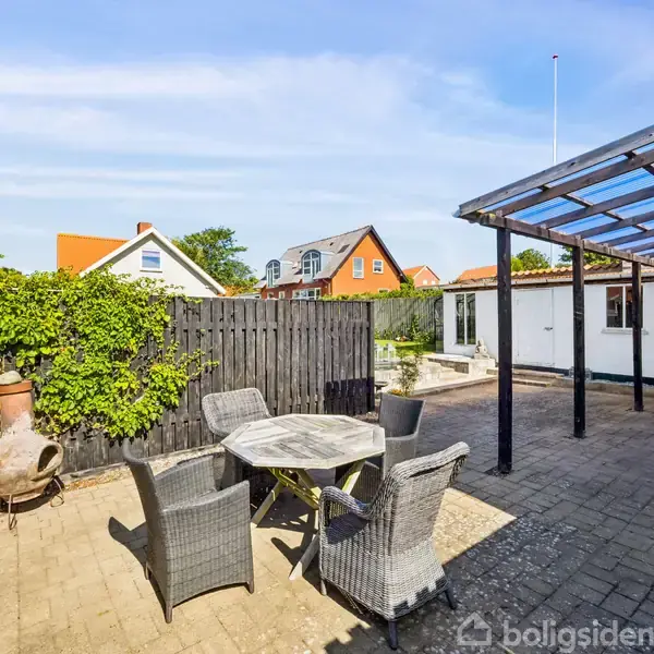 Rundt bord med fire kurvemøbler står på en flisebelagt terrasse. Til højre et halvtag, og til venstre en træhegn med planter. Røde hustage er synlige i baggrunden.