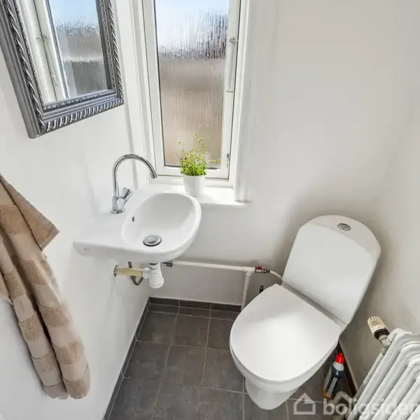 Et lille badeværelse med et toilet ved siden af en hvid håndvask. Et lille vindue med en plante står over vasken, og et håndklæde hænger på væggen.