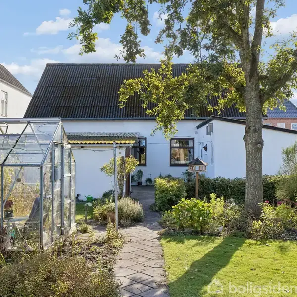 Et hvidt hus med sort tag står i en frodig have. Et drivhus er synligt til venstre, og stier fører gennem blomsterbede. Træer og buske omkranser området.