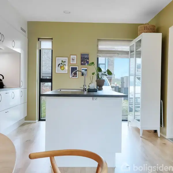 Et moderne køkken med en hvid køkkenø fyldt med planter. Overskabe og skuffer er hvide, en elkedel står på en sort køkkenbordplade. Grønne vægge med kunst på væggen.