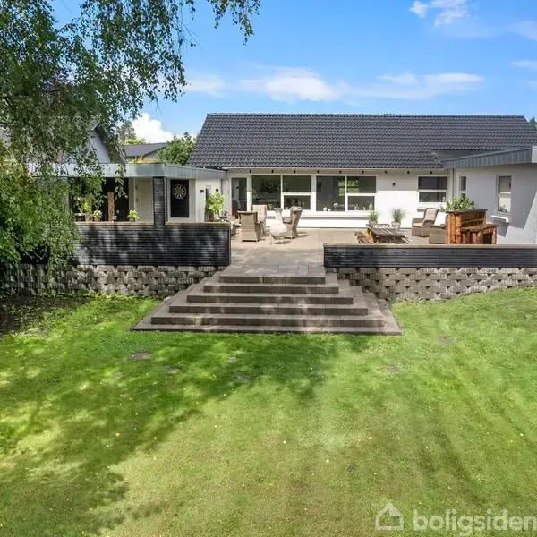 Et moderne hus med grå tagsten står foran en stor terrasse med havemøbler. Omkringliggende grøn græsplæne og træer skaber en fredelig haveatmosfære under en klar, blå himmel.
