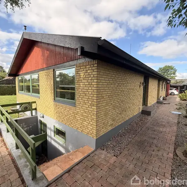 Et hus af gule mursten med et skråt tag står i en velplejet have. En sti af stenfliser fører forbi huset omgivet af græsplæne og buske med blå himmel i baggrunden.