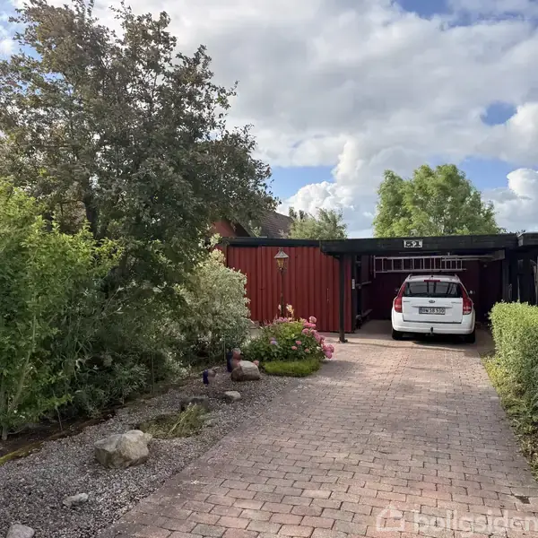 En hvid bil holder parkeret i en indkørsel foran en rød bygning med garage. Omkring er der grønne buske og træer under en delvist skyet himmel. Husnummeret "63" ses over garagen.