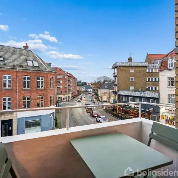 En lille balkon med bord og stole står åben mod en byudsigt, hvor røde murstensbygninger og butikker strækker sig langs en vej under en klar blå himmel.