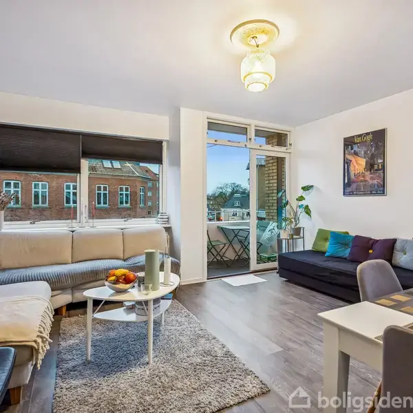 En moderne stue har en lys sofa og sofabord med frugtskål. Stuen fører ud til en balkon med et lille bord og stole, omgivet af bygninger udenfor.