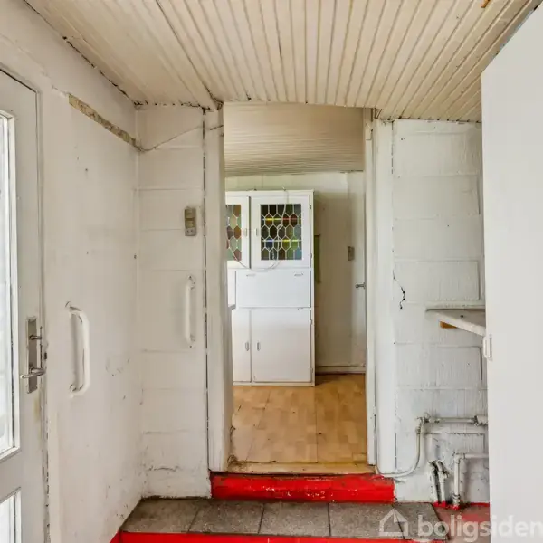 Dør indgangen fører til en gang med hvidmalede vægge, rødt gulvtæppe og en glasdør til venstre. I bunden ses en reol med farvet glas i et lyst rum.